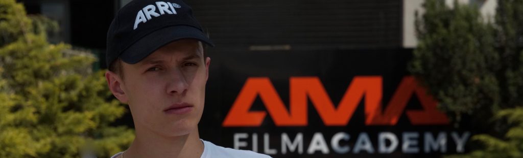 Młody mężczyzna w czapce przed znakiem szkoły filmowej AMA