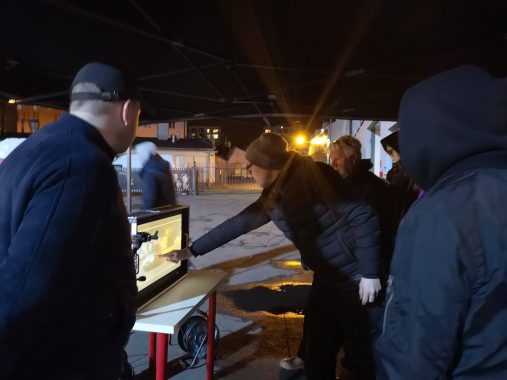 Grupa osób oglądająca zawartość ekranu monitora nocą na zewnątrz