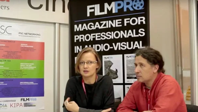 Dwie osoby rozmawiają przy stoisku FILMPRO, magazynu dla profesjonalistów audiowizualnych