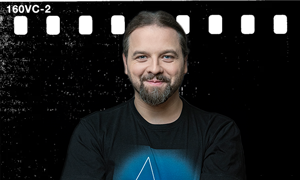 Mężczyzna z brodą w czarnej koszulce z niebieskim logo na tle filmu