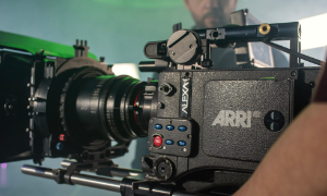 Zbliżenie na profesjonalną kamerę filmową ARRI ALEXA na planie filmowym, a w tle specjalista ds. kamer niestałych z Krakowa reguluje sprzęt. Kamera jest zamontowana na statywie, a oświetlenie studyjne tworzy kinową atmosferę.