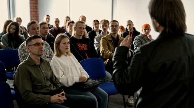 Grupa studentów siedzi na niebieskich krzesłach słuchając wykładu podczas dnia otwartego w szkole filmowej AMA