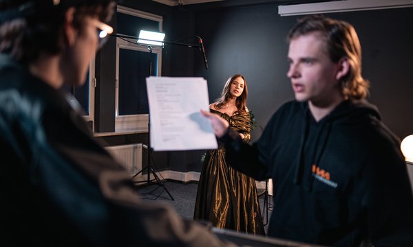 Kobieta w sukni stoi przed światłami studyjnymi, podczas gdy dwie osoby na pierwszym planie omawiają lub przeglądają scenariusz – jedna z nich trzyma kartkę, a druga ma na sobie okulary.